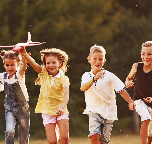 Fünf Kinder rennen bei Sonnenschein über eine Wiese. Das mittlere Kind hält dabei ein Modellflugzeug in der Hand.