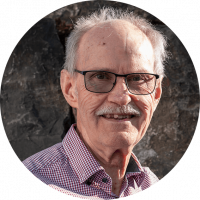 Mann mit grauen Haaren, Schnurrbart und Brille steht vor einer Steinwand. Man sieht nur den Kopf bis zum Hals.