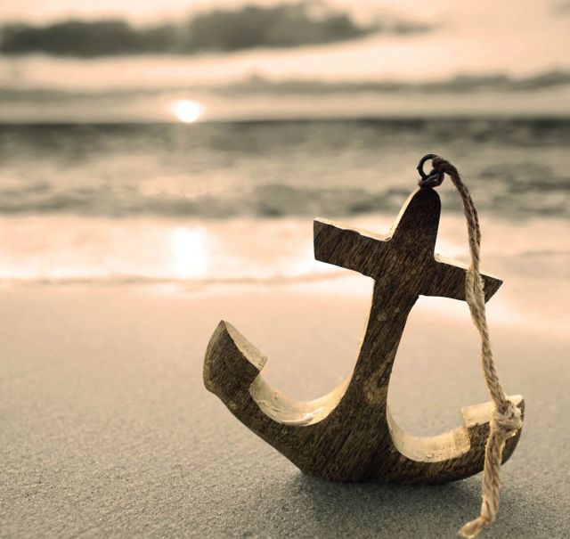 Kleiner Miniatur-Holzanker mit Schnur obendran steht im Sand. Im Hintergrund sieht man das Meer und die untergehende Sonne.