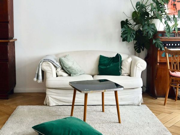 Auf hellem Parkettboden steht an einer weißen Wand ein weißes Sofa für 2 Personen. Vor dem Sofa liegt ein weißer Teppich, auf dem Teppich steht ein Hocker. Vor dem Hocker liegen 2 dunkelgrüne Kissen.