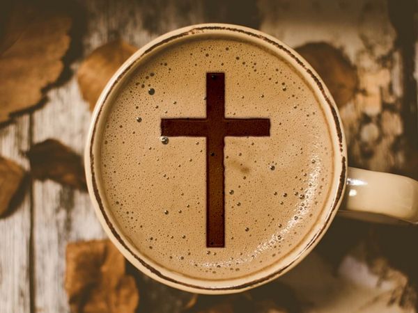 Eine Tasse Kaffee steht auf einem Tisch mit Herbstblätter dekoriert. Im weißen Michschaum ist ein braunes Kreuz,