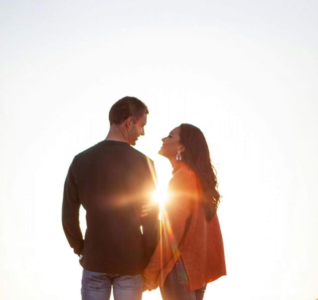Ein junges Paar steht auf einer Wiese vor einem Berg und die Sonnenstrahlen  strahlen zwischen ihnen durch.