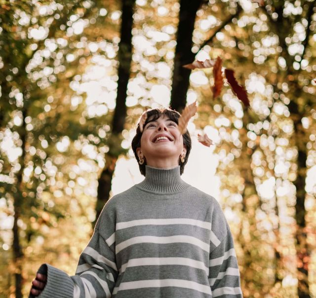 Eine Frau steht mit grauem Pulli in einem herbstlichen Wald und schaut nach oben, während rote Blätter von den Bäumen fallen.