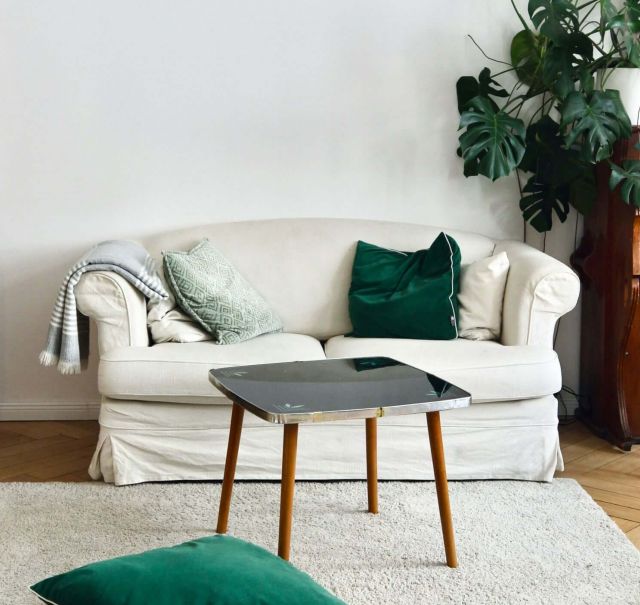 Auf hellem Parkettboden steht an einer weißen Wand ein weißes Sofa für 2 Personen. Vor dem Sofa liegt ein weißer Teppich, auf dem Teppich steht ein Hocker. Vor dem Hocker liegen 2 dunkelgrüne Kissen.