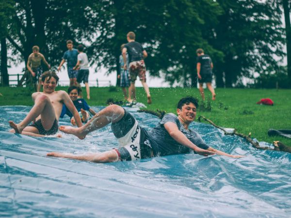 Jungs rutschen in Badehose über eine mit Wasser geflutete Plane einen Hügel herunter.