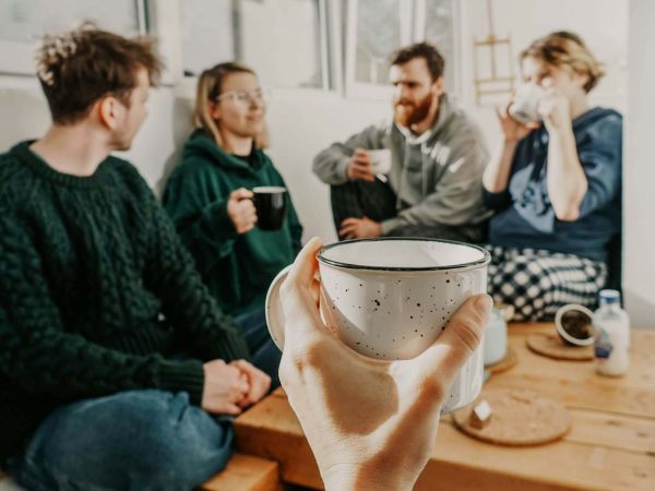 4 Personen sitzen auf dem Boden in einer Zimmerecke und trinken Kaffee. Vor den Personen hält jemand aus Kameraperspektive eine weitere Kaffeetasse ins Bild.