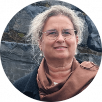 Frau mit blonden Haaren, Brille und braunem Schal steht vor einer Steinwand. Man sieht nur den Kopf bis zum Hals.