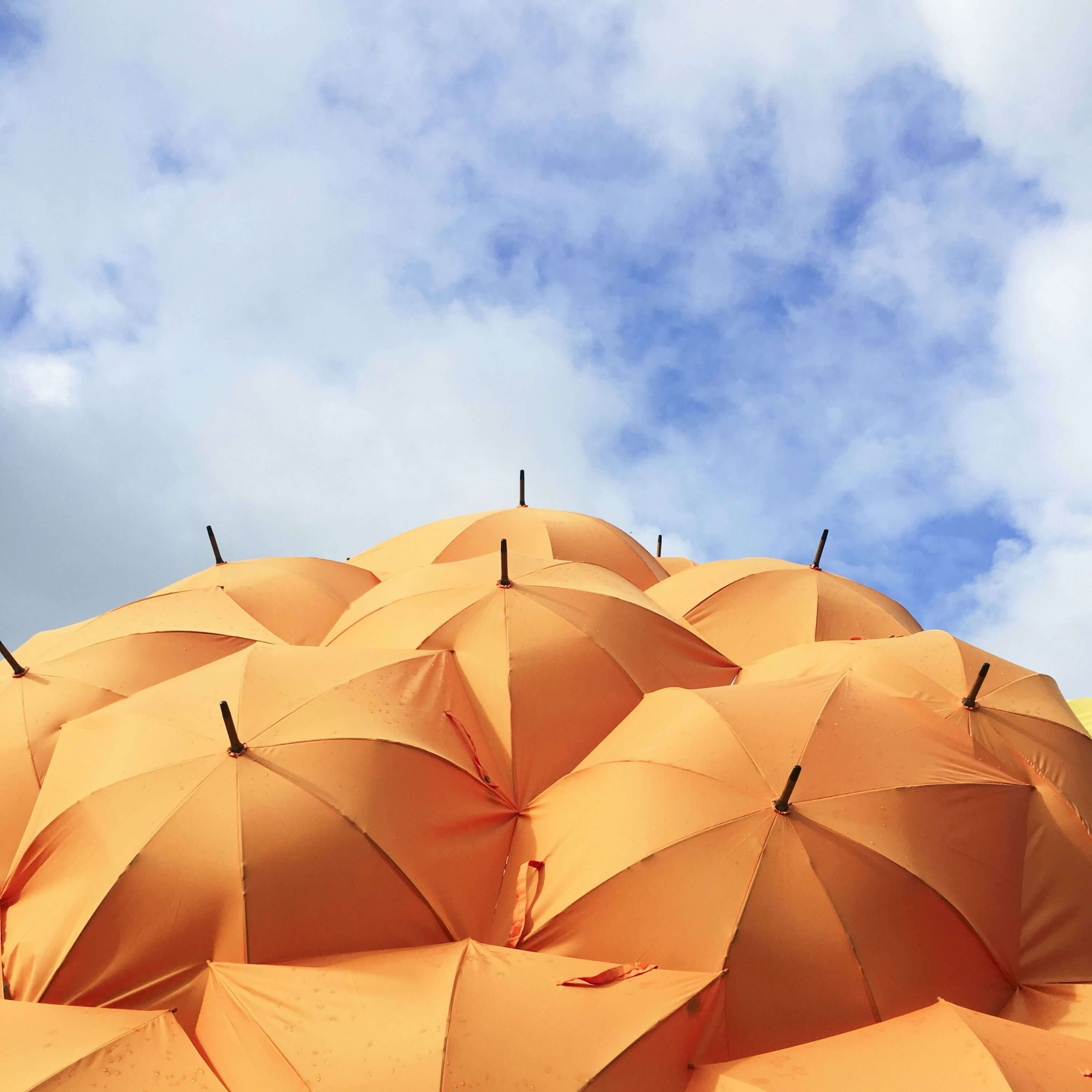 Oben auf dem Bild sieht man blauen Himmel mit leichten Wolken und unten auf dem Bild einen Haufen mit orangenen aufgespannten Regenschirmen.