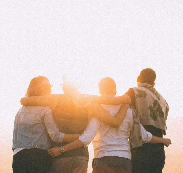 4 Jugendliche stehen in einer Reihe auf einem Berg und lege sich die Arme über die Schultern. Sie sind mit dem Rücken zu sehen und schauen sich den Sonnenuntergang an.
