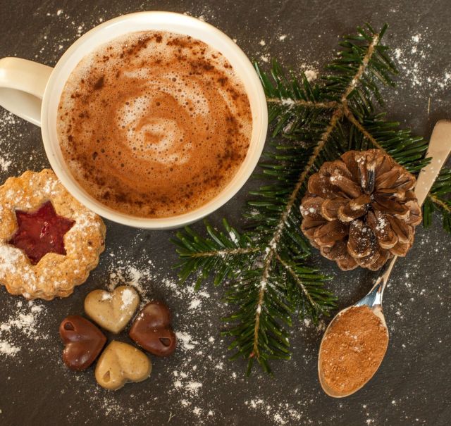 Auf einem Tisch steht eine weiße Tasse mit Kaffe gefüllt, auf dem Kaffee ist weißer Milchschaum. Um die Tasse herum liegen gepuderte Kekse, ein grüner Tannenzweig, ein Tannenzapfen und ein kleiner Löffel  mit Zimt.