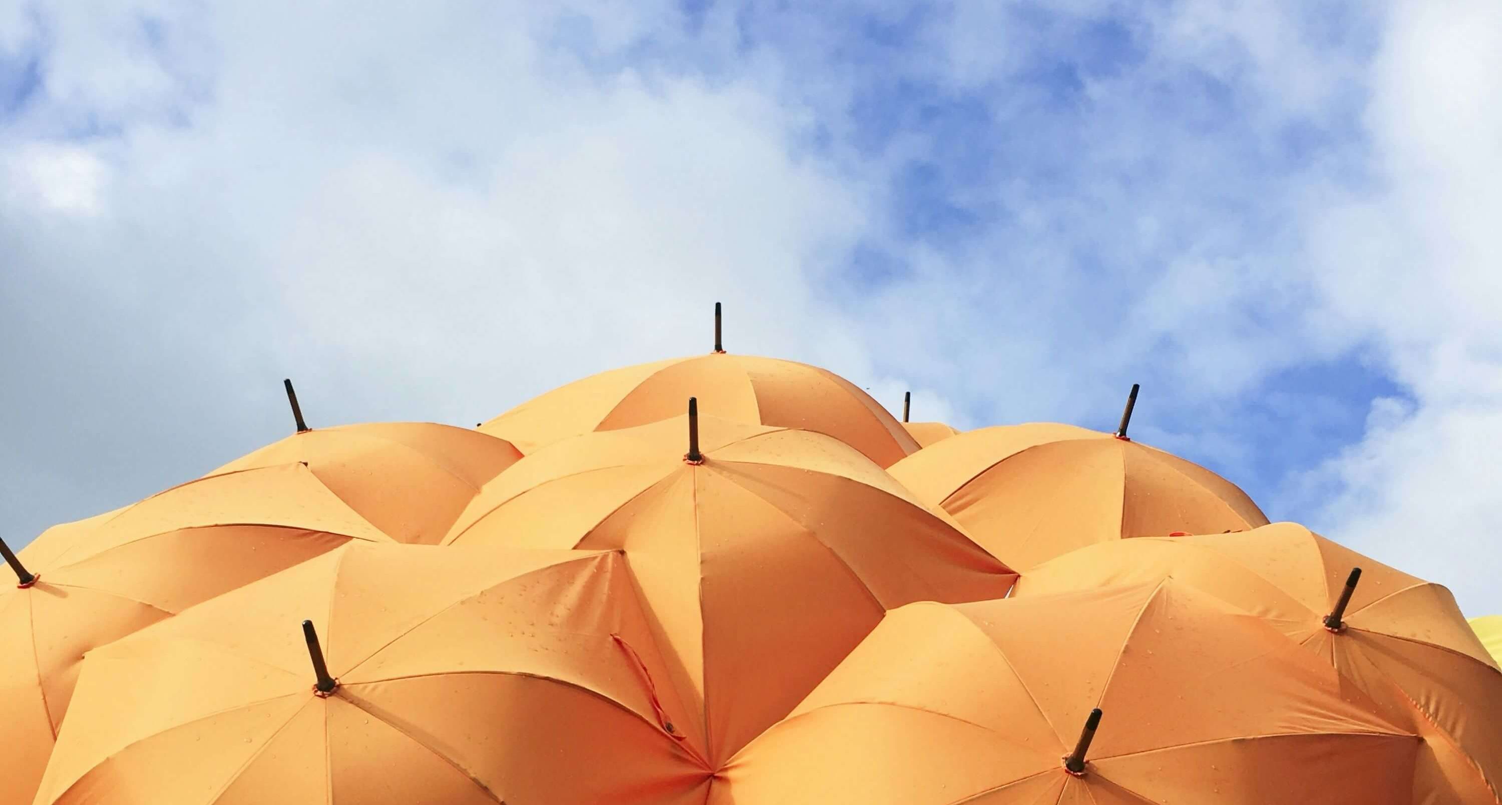 Oben auf dem Bild sieht man blauen Himmel mit leichten Wolken und unten auf dem Bild einen Haufen mit orangenen aufgespannten Regenschirmen.