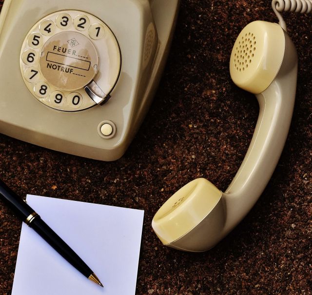 Ein altes, beiges Telefon mit Wählscheibe steht auf braunem Untergrund. Der Hörer liegt daneben. Rechts daneben  steht  ein Zettelkasten. Ein  Zettel liegt vor dem Telefon. Auf dem Zettel liegt ein schwarz goldener Kugelschreiber.