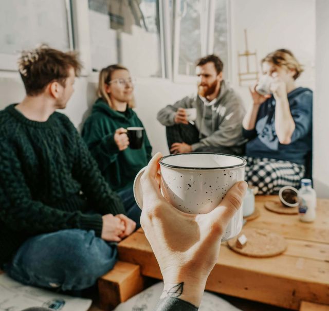 4 Personen sitzen auf dem Boden in einer Zimmerecke und trinken Kaffee. Vor den Personen hält jemand aus Kameraperspektive eine weitere Kaffeetasse ins Bild.