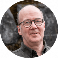 Mann mit grauen Haaren und Halbglatze steht vor einer Steinwand. Er trägt eine Brille. Man sieht nur den Kopf bis zum Hals.