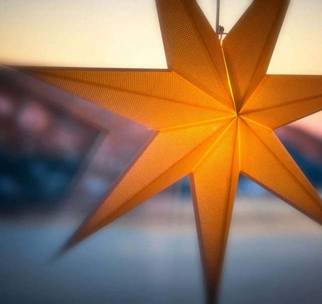 Ein großer gelber Faltstern hängt vor einem Fenster. Im Hintergrund sieht man einen Sonnenuntergang.