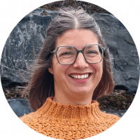 Frau mit braunen Haaren, Brille und orangenem gestricktem Pullover steht vor einer Steinwand. Man sieht nur den Kopf bis zum Hals.