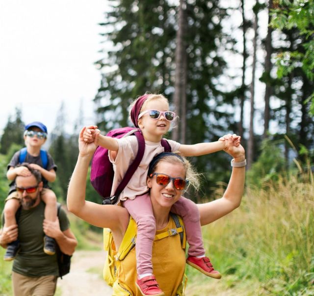 4-köpfige Familie im Wald. Die Kinder sitzen auf den Schultern der Eltern. Alle tragen Sonnenbrillen.