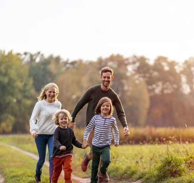 Eine Familie (bestehend aus Vater, Mutter und 2 Kindern) läuft gemeinsam über einen Feldweg und lacht.