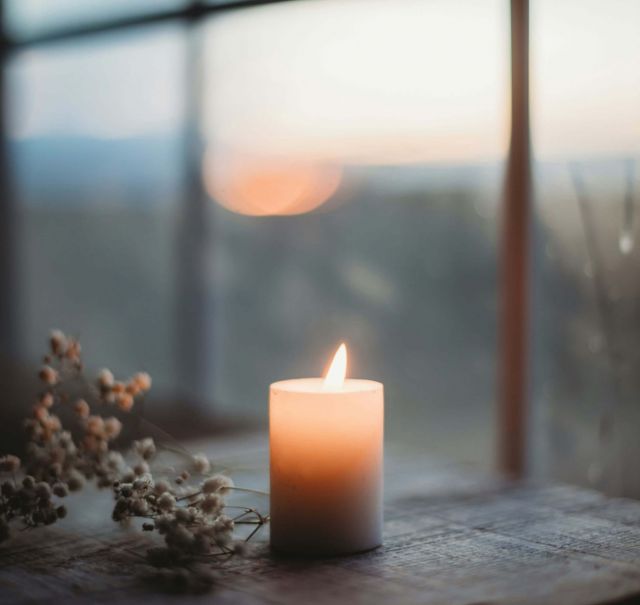 Eine angezündete Kerze steht innen vor einem Sprossenfenster. Durch das Fenster sieht man den Sonnenuntergang.