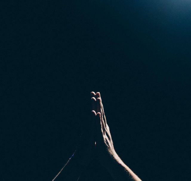 Zwei Hände sind mit den Handflächen aneinander nach oben gerichtet (wie bei einem Gebet) und werden vom Licht angestrahlt. Der Hintergrund ist schwarz.