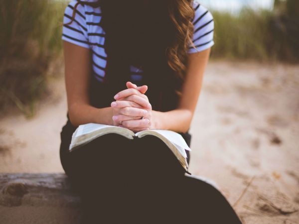 Eine Frau sitzt im Sand auf einem Stein. Sie hat eine Bibel auf Ihrem Schoß und die gefalteten Hände liegen auf dem Buch. Ihr Kopf ist nach unten geneigt.