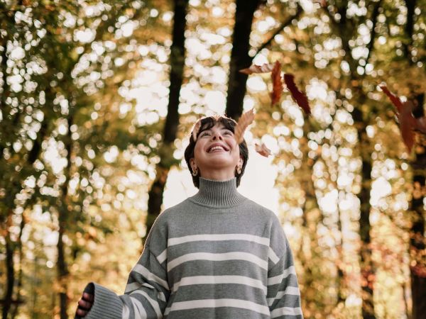 Eine Frau steht mit grauem Pulli in einem herbstlichen Wald und schaut nach oben, während rote Blätter von den Bäumen fallen.