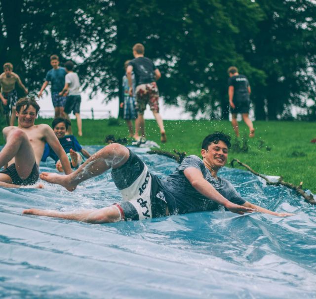 Jungs rutschen in Badehose über eine mit Wasser geflutete Plane einen Hügel herunter.