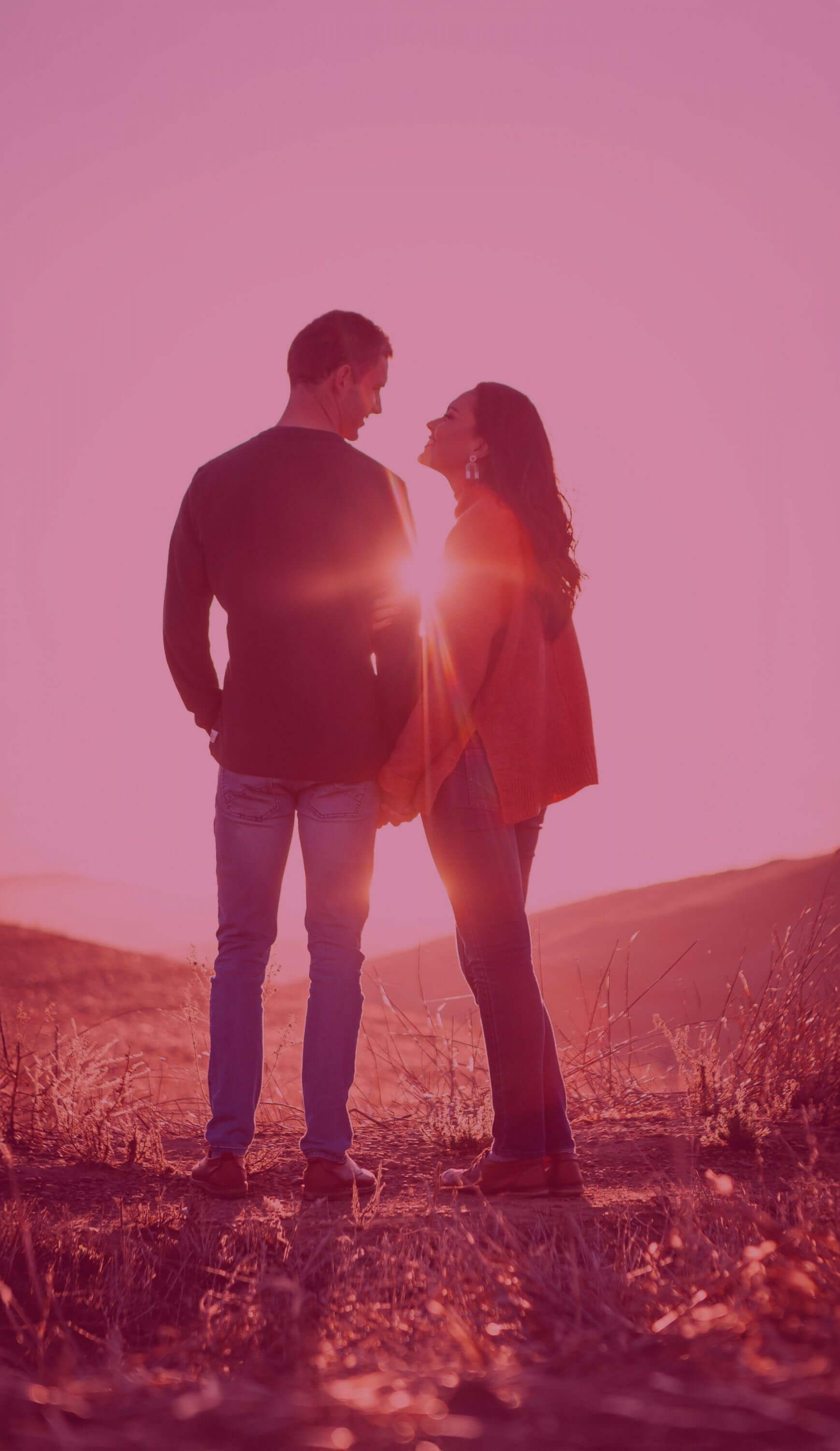 Ein junges Paar steht auf einer Wiese vor einem Berg und die Sonnenstrahlen strahlen zwischen ihnen durch.