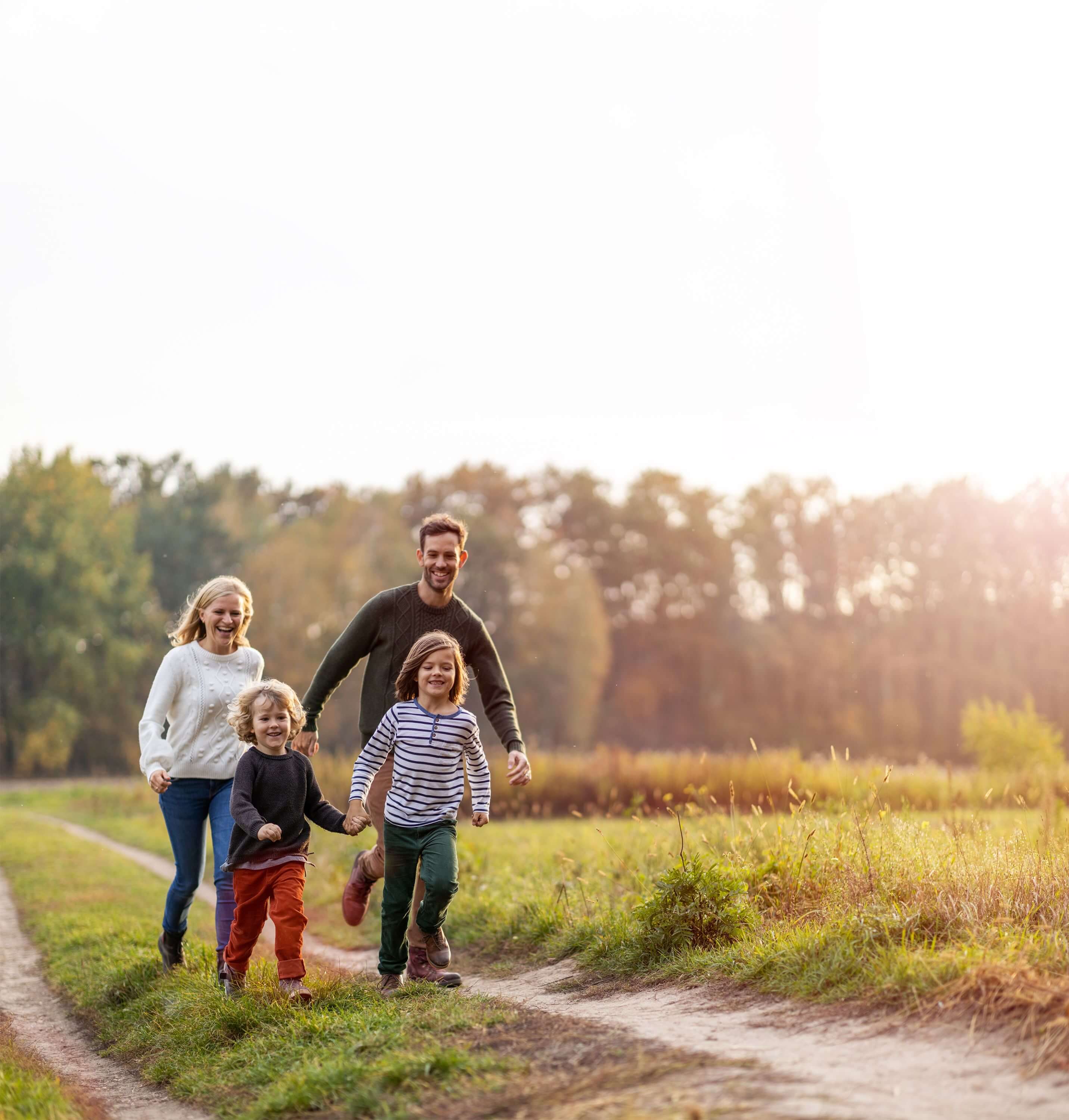Eine Familie (bestehend aus Vater, Mutter und 2 Kindern) läuft gemeinsam über einen Feldweg und lacht.