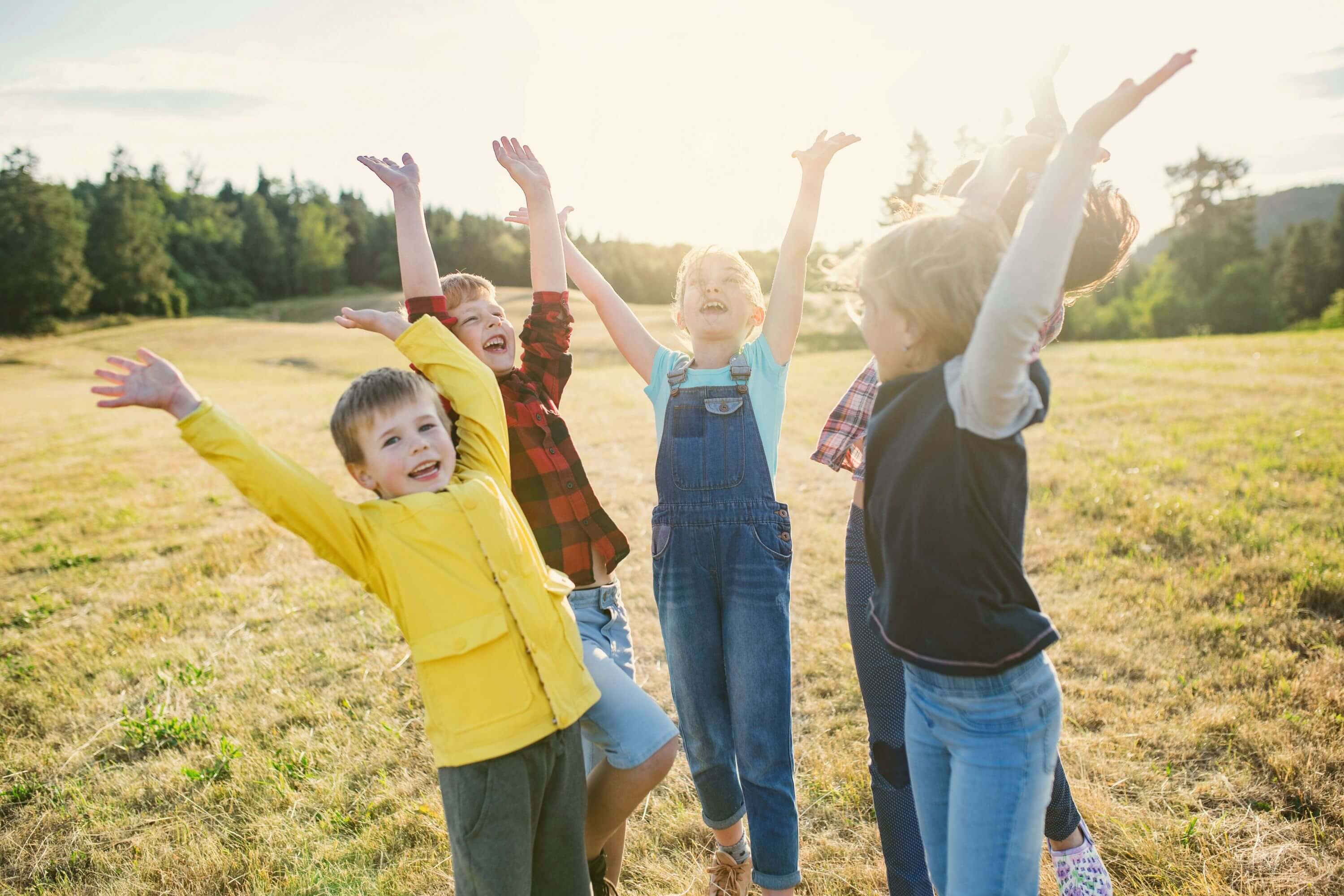5 Kinder stehen auf einer Wiese und strecken ihre Arme fröhlich in den Himmel.  Im Hintergrund sieht man grünen Wald