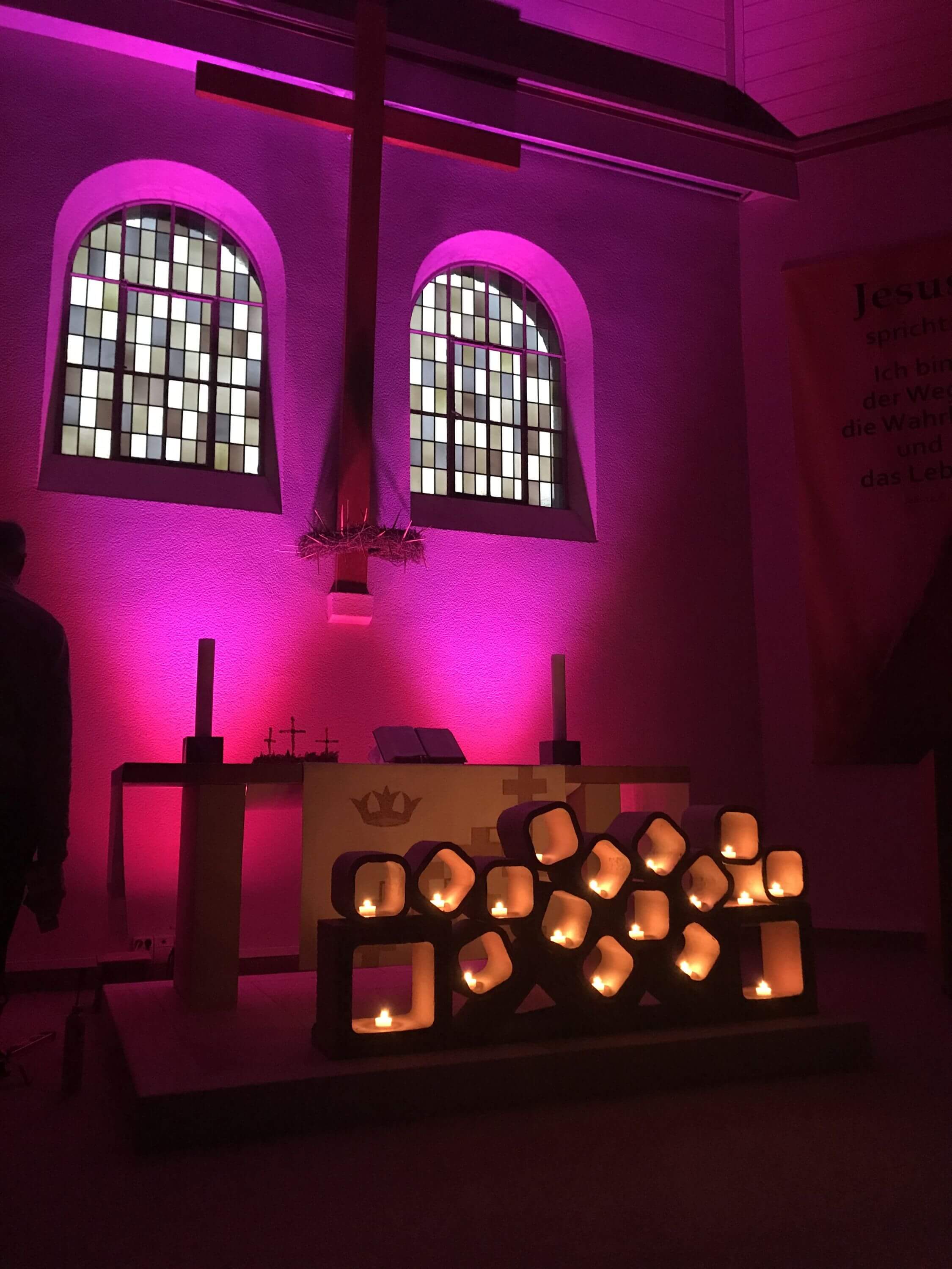 Eine Kirche mit Glasbausteinfenstern. Ein großes Holzkreuz hängt zwischen den Fenstern und wird mit lila Lich angestrahlt. Vor dem Kreuz steht ein Altar. Vor dem Altar sind kleine Kästchen aufeinandergestellt. In den Kästchen sind Lichter.