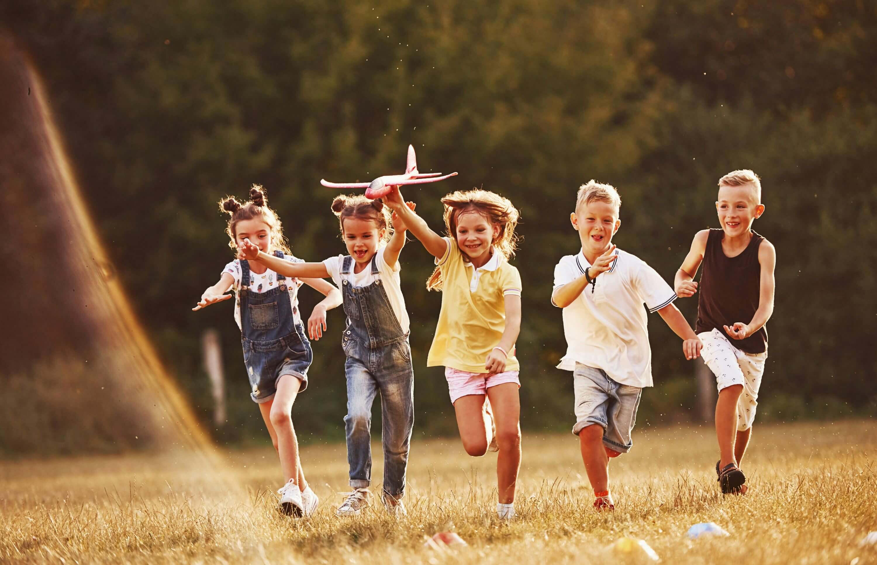 Fünf Kinder rennen bei Sonnenschein über eine Wiese. Das mittlere Kind hält dabei ein Modellflugzeug in der Hand.