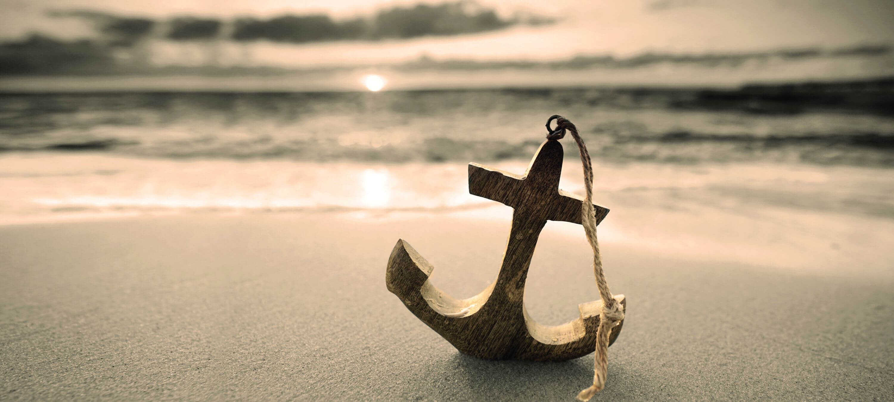 Kleiner Miniatur-Holzanker mit Schnur obendran steht im Sand. Im Hintergrund sieht man das Meer und die untergehende Sonne.